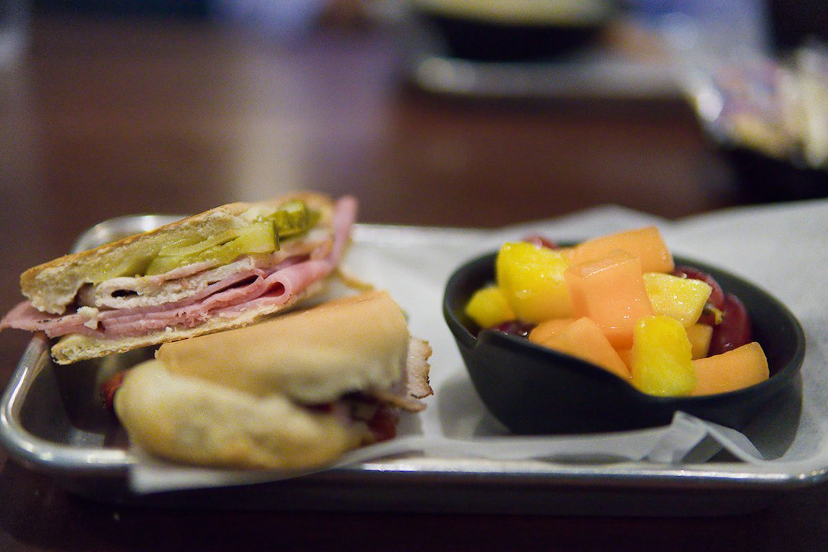 Pressed ham sandwich with pickles next to a bowl of fresh fruit